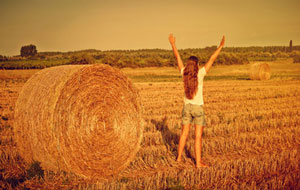 barefoot woman in open field standing next to hay with arms lifted up high