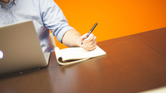 Guy sitting by a laptop writing something down.