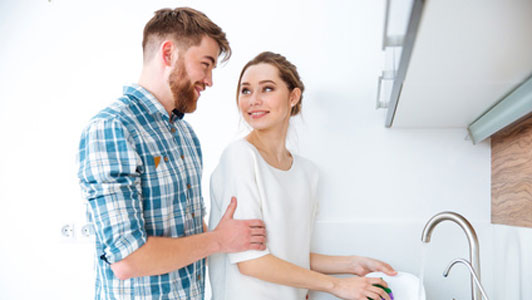 man standing behind woman doing dishes