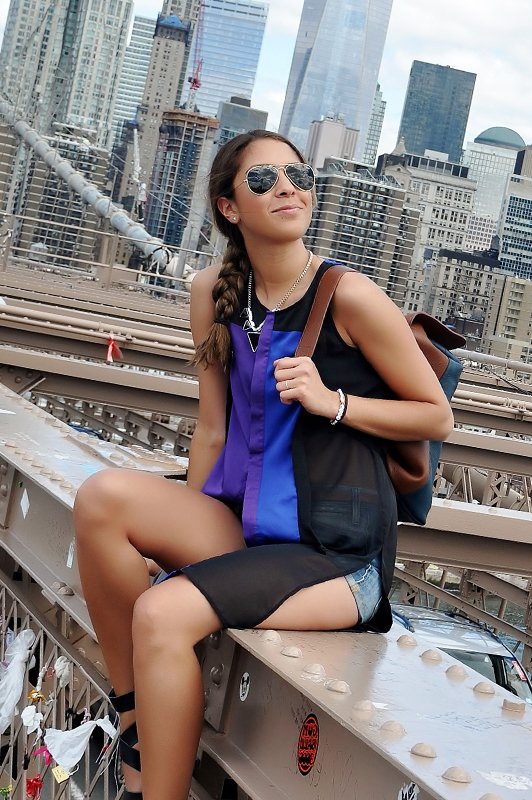 Girl wearng sunglasses and shorts holding a backpack while sitting outside.