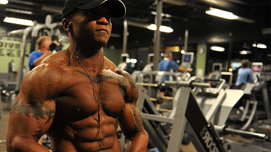 Muscular tattooed guy in a gym wearing a black hat on his head.