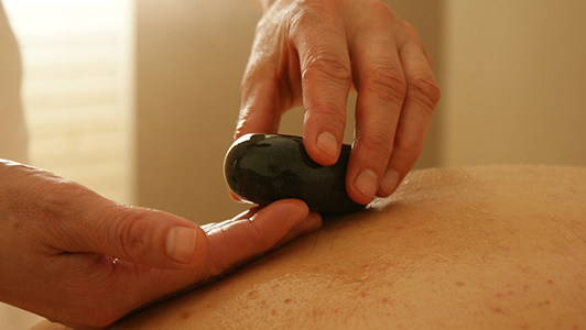 Hands holding a black stone used for massage over someone's back.