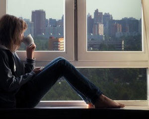 woman sitting on window sill reflecting on view of city drinking a cup of coffee