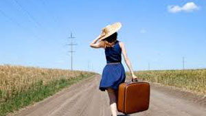 woman in blue dress carrying luggage walking down road into the distance 