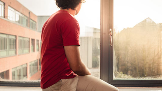 young man with curly hair wearing red shirt sitting on window sill looking outside alone