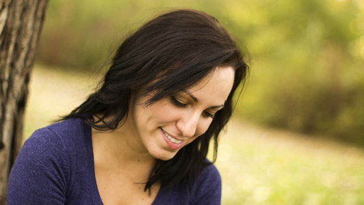 Brunette in a violet blouse sitting by a tree and smiling.