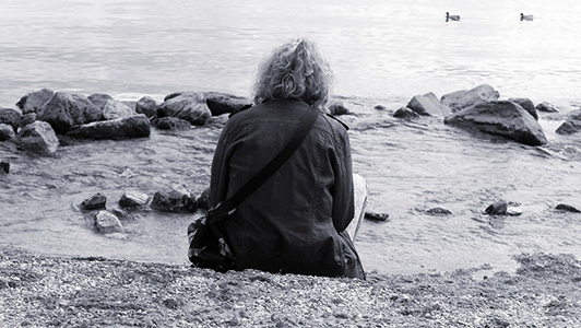 Guy with long curly hair wearing a jacket sitting near a lake.