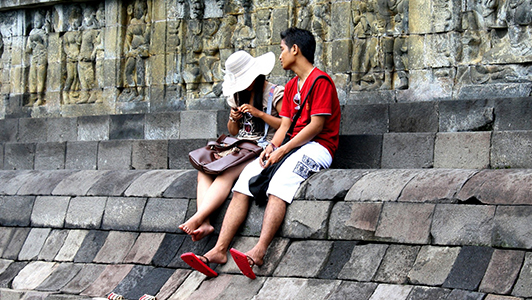 Girl with a hat sitting on a wall with a guy in red tee shirt.