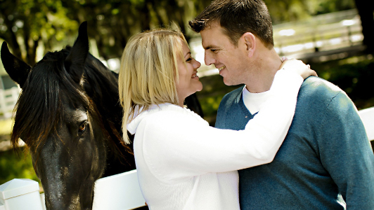 A couple smiling and looking at each other while standing next to a horse.