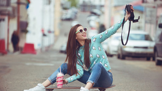 young woman wearing sunglasses sitting on skateboard drinking shake taking picture of self