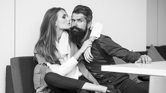 black and white photo of bearded man with arm of woman around him on couch in front of table