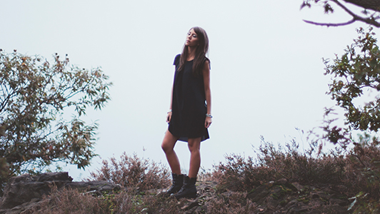 Girl in black dress and boots standing near a bush.