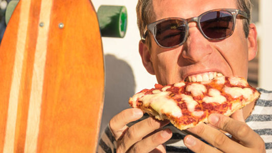 man in sunglasses next to skateboard eating a slice of pizza