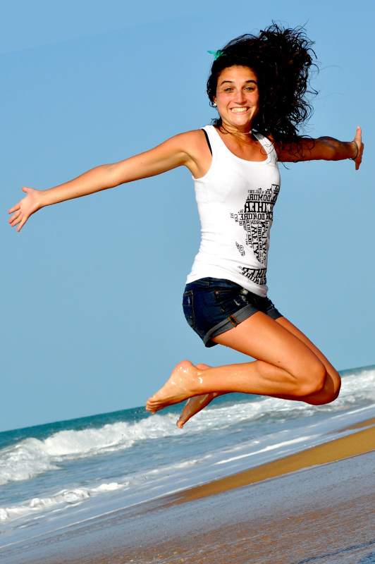 Girl on a beach wearing denim shorts and white top while jumping.