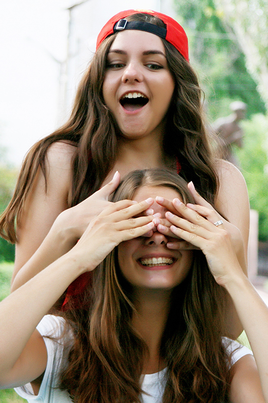 Two girl with long hair, one covering the eyes of the other.