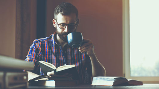 guy with beard with glasses drinking coffee and reading a book at desk